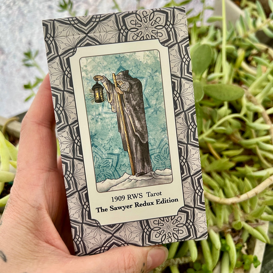 Hand holding a tarot card book titled '1909 RWS Tarot The Sawyer Redux Edition' with a decorative cover against a plant background.
