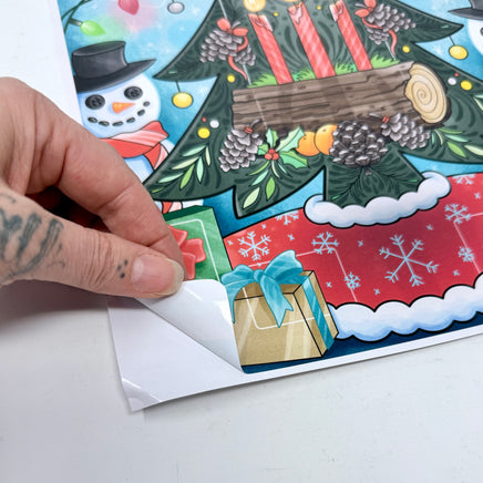 Hand peeling the corner on a Christmas-themed sticker with a snowman and festive decorations.