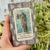 Hand holding a tarot card book titled '1909 RWS Tarot The Sawyer Redux Edition' with a decorative cover against a plant background.