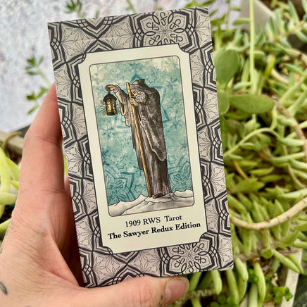 Hand holding a tarot card book titled '1909 RWS Tarot The Sawyer Redux Edition' with a decorative cover against a plant background.